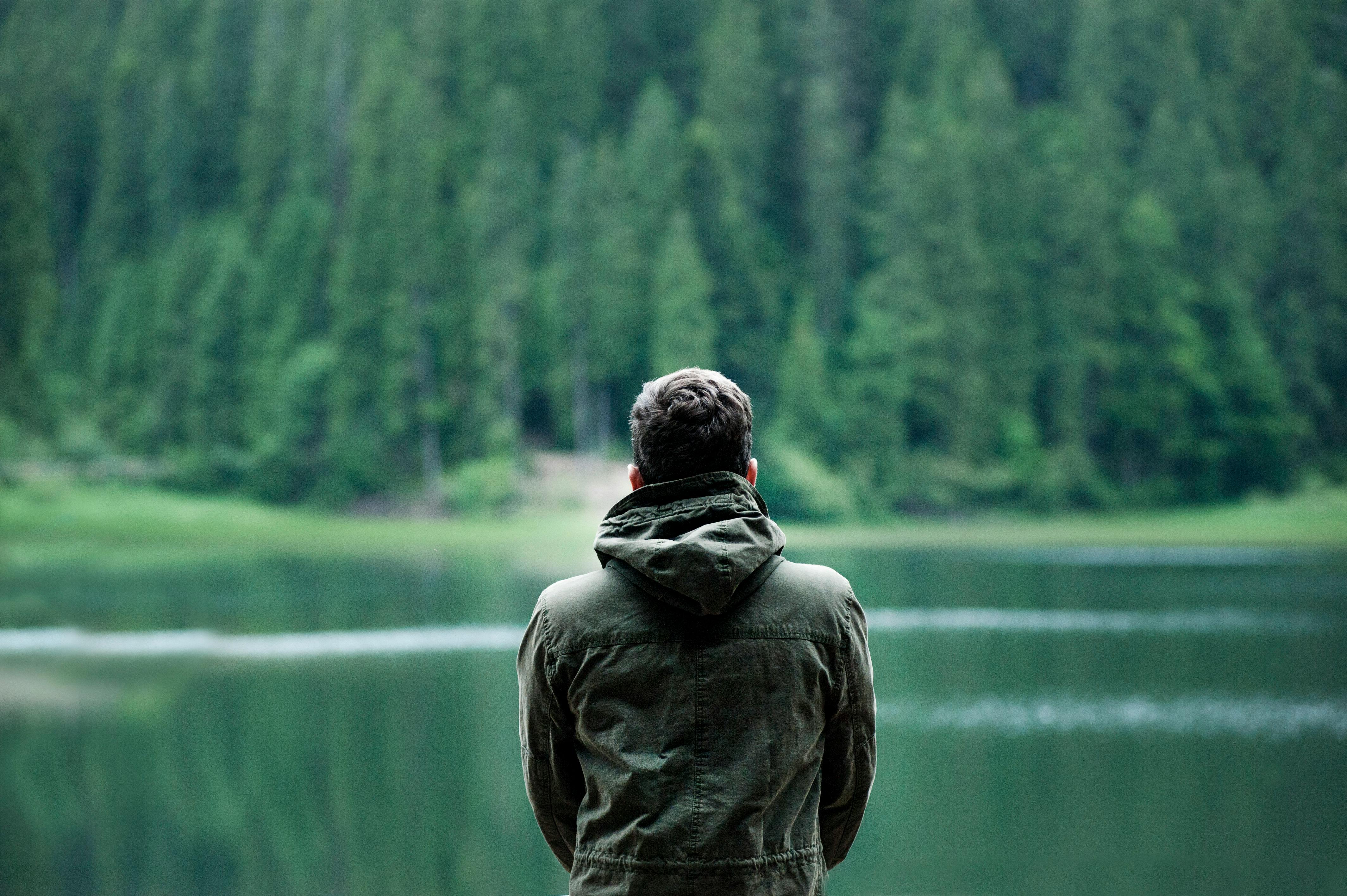 a man wearing a green coat looking out to a lake and a forest